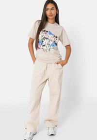 Beige grafische t-shirt met bloemenontwerp, korte mouwen, relaxte pasvorm; gecombineerd met lichte beige losse broeken en witte sneakers met grijze accenten.
