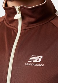 Hnědá bunda na zip s vysokým límcem, s jasně béžovým zipem a vyšívaným bílým logem. Vyrobena z texturované látky.