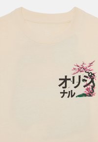 Beige T-Shirt mit kurzen Ärmeln, auf der linken Seite mit stilisiertem, schwarzem Text, einem pinken Kirschblüten-Design und grünen Akzenten.