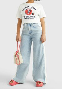 Zkrácená bílá tričko s grafickým potiskem, světle modré široké džíny, růžové sandály a béžová taška na šňůrku s růžovými akcenty.