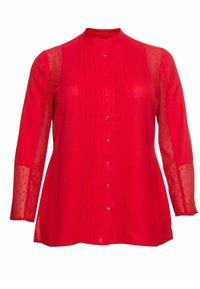 Blouse rouge avec col montant, manches longues en voile transparent à pois, boutonnée sur le devant, et plis verticaux sur le corsage. Tissu doux et léger.