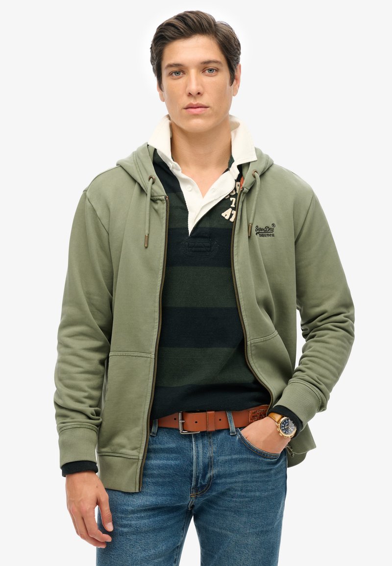 Sudadera con cremallera de color verde oliva y cordones, llevada sobre una camisa polo a rayas negras y verdes. Unos vaqueros de mezclilla y un cinturón marrón completan el look.