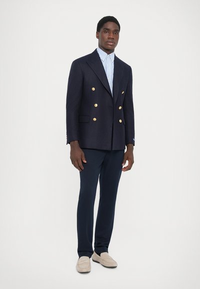 Blazer double boutonnage bleu marine foncé avec des boutons dorés, associé à une chemise rayée bleu clair et un pantalon navy, complété par des mocassins beiges.