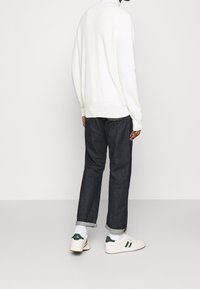 Weißer hoher Pullover, dunkelblaue Jeans und weiße Sneakers mit grünen Akzenten. Lockere Passform, umgeschlagene Bündchen an den Jeans, minimalistisches Design.