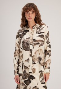 Blouse à motif floral en crème avec de grands motifs de fleurs brunes et noires. Dispose d'un col, d'une fermeture à boutons et de manches longues. Texture lisse.