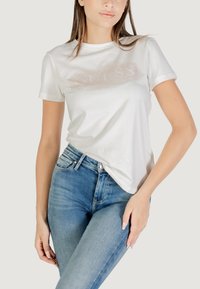 Camiseta de algodón blanca de manga corta, con un diseño brillante de logo "GUESS" en la parte frontal. Combinada con unos jeans de tiro alto azules.