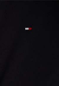 Svart tyg med en texturerad väv som har en liten broderad logotyp: en horisontell rektangel med röd, vit och blåa ränder.