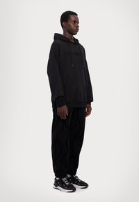 Sudadera de algodón negra con un bolsillo de canguro frontal y capucha con cordón, combinada con pantalones negros de patrón y zapatillas negras.