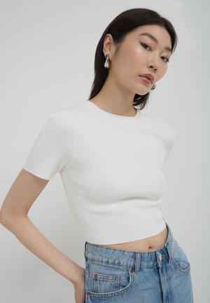 Jeune femme aux longs cheveux noirs portant un top court à manches courtes blanc, un jean taille haute bleu et des boucles d'oreilles argentées, posant devant un fond uni.