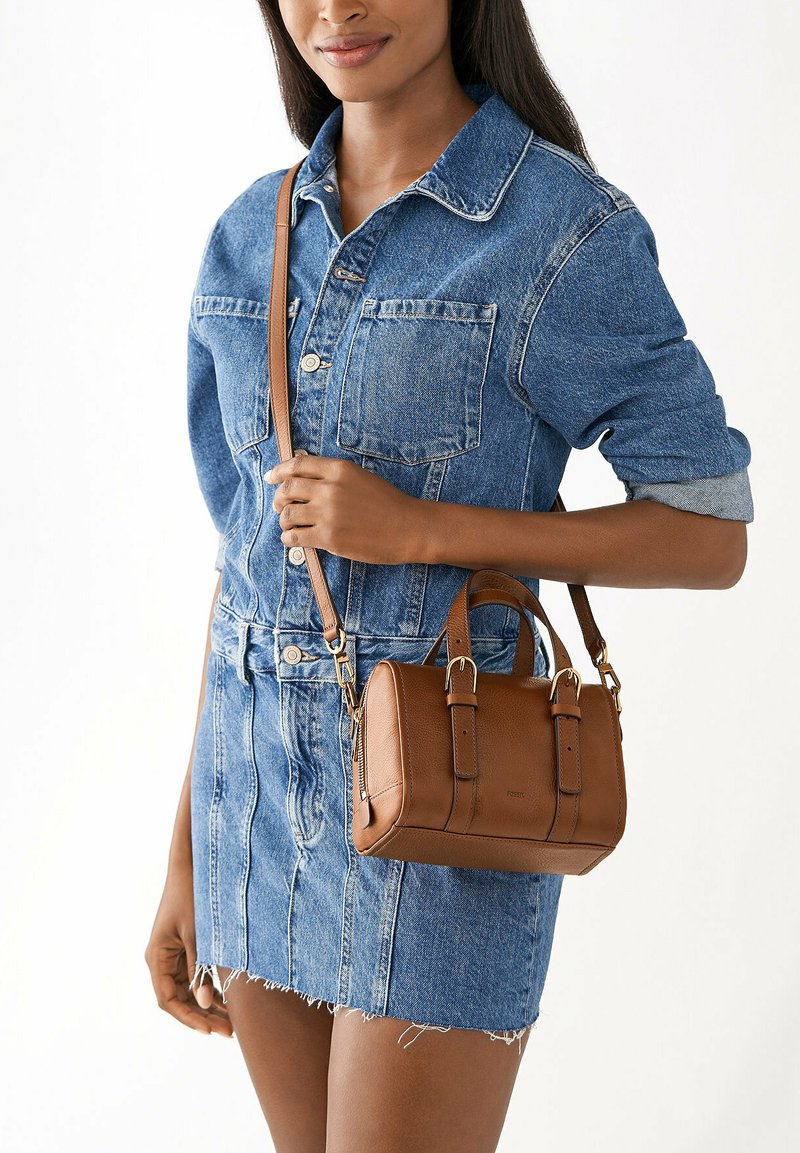Sac à main en cuir marron avec double anses et une bandoulière amovible, présentant une texture lisse et un minimum de matériel. Porté avec une veste en denim et une jupe.