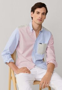 Jeune homme assis sur une chaise en bois portant une chemise à manches longues avec des rayures verticales rouges, bleues et beiges, et un pantalon blanc, sur un fond uni.