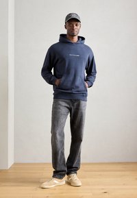 Hanorac din bumbac navy cu buzunar frontal, având un logo alb. Purtați-l cu blugi gri și adidași bej pe un podea din lemn deschis.