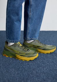 Grüne Sportschuhe mit gelben Akzenten, Obermaterial aus Mesh, strukturierten Details und gepolstertem Kragen, vor blauer Teppich und Jeans.