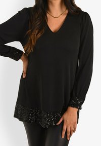 Black long-sleeve top with a V-neck, featuring textured sequin accents on the sleeves and hem, made from a smooth, flowing fabric.
