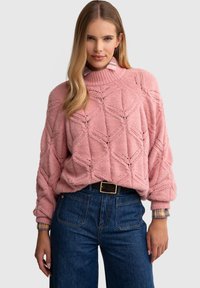 Roosa kudumine sviiter, millel on geomeetriline muster ja ribakrae; paaritatud kõrge vöökohaga tumedate teksadega, millel on ees taskud.