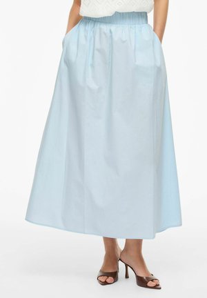 Femme portant une jupe midi bleu clair avec une taille élastique et des poches, associée à des sandales à talons hauts ouvertes marron.