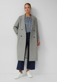 Grauer doppeltknöpfender Mantel mit maßgeschneidertem Design, kombiniert mit einem gemusterten Oberteil, dunklen weit geschnittenen Jeans und weißen Sneakers.