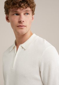 Fehér pólóing, textúrált kötött mintázattal és cipzáras záródással. Klasszikus gallérral rendelkezik, és testszerváltó a vállaknál és a mellkason.