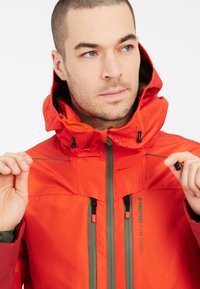 Helle orange wasserdichte Jacke mit Kapuze, versehen mit grauen Akzenten, Belüftungsöffnungen und verstellbaren Kordeln am Kragen.