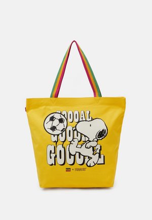 Bolsa de mano amarilla con un personaje de dibujos animados pateando un balón de fútbol. Presenta asas de rayas coloridas y texto en gran tamaño. Diseño de tela duradera.