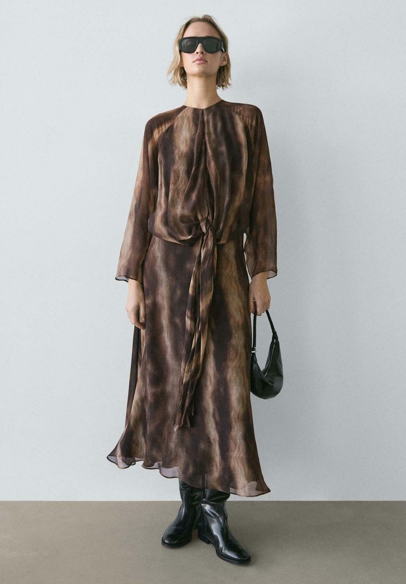 Robe longue marron à motifs avec détail noué à la taille, manches longues en voile transparent, et silhouette fluide. Associée à des bottes noires montantes jusqu'aux genoux et un sac.