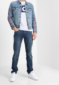 Světlě modrá džínová bunda s červenými a bílými pruhovanými doplňky, šedé tričko s kulatou grafikou, tmavě modré džíny a bílé tenisky.