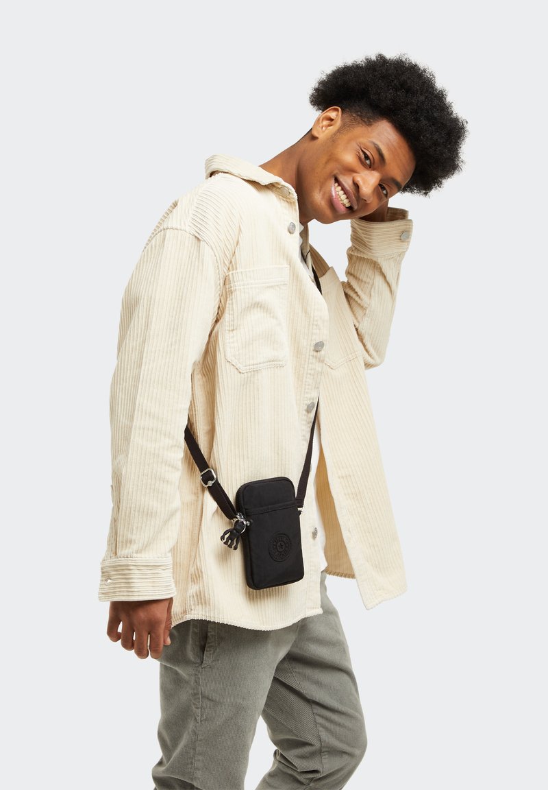 Custodia per telefono in tessuto nero con zip, tracolla regolabile e dettaglio del logo, indossata sopra una camicia in corduroy chiaro e pantaloni verdi.