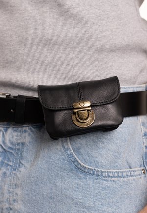 Pochette en cuir noir avec rabat incurvé et fermoir en laiton antique, attachée à une ceinture noire, positionnée près d'un jean en denim bleu.