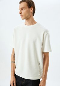 Hvid bomulds t-shirt med rund halsudskæring, korte ærmer og en sort tekstdetalje ved kanten, parret med mørke jeans.