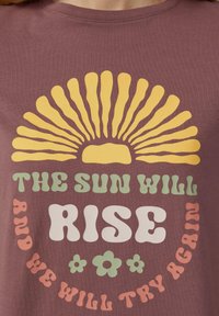 T-shirt en coton marron avec un grand motif de soleil en jaune et un texte en couleurs variées : "LE SOLEIL SE RÉVEILLERA ET NOUS ESSAYERONS ENCORE."