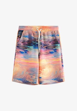 Teenagedreng står iført en matchende hættetrøje og shorts med et flerfarvet abstrakt mønster samt hvide sokker og sneakers mod en ensfarvet baggrund.