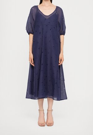 Frau trägt ein marineblaues Midikleid mit transparenten Puffärmeln und strukturiertem Stoff, kombiniert mit nudefarbenen Sandalen mit Absatz und Knöchelriemen.