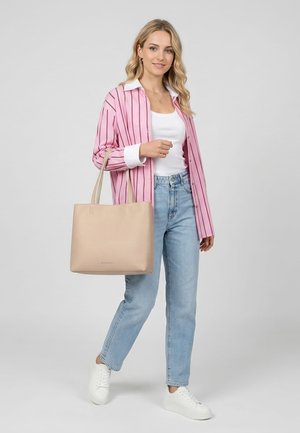 Jauna sieviete rozā svītrainā krekliņā, baltā topā, zilās džinsu biksēs, baltās sporta kurpēs, rokās tur pelēkā fonā bēša krāsas plecu somu.