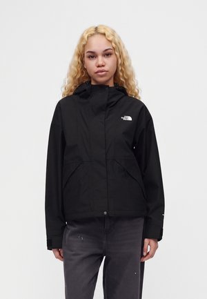 Mladá osoba s dlhými kučeravými blond vlasmi, oblečená v čiernu bundu North Face a tmavé rifle, stojaca pred jednoduchým svetlým pozadím.
