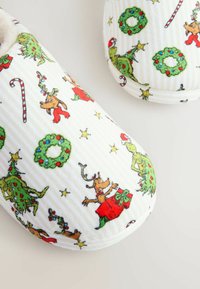 Pantofole bianche e soffici con un motivo natalizio che presenta elementi festivi come alberi, ghirlande, bastoncini di zucchero e personaggi. Superficie textured.