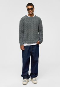 Grijze kabeltrui met een ronde halslijn, gedragen over een wit T-shirt. Gecombineerd met losse spijkerbroek en lichtgekleurde sneakers.