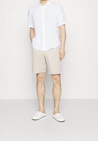 Camicia bianca in lino a maniche corte, pantaloni beige e scarpe slip-on bianche. Abbigliamento casual con una vestibilità rilassata e dettagli minimi.