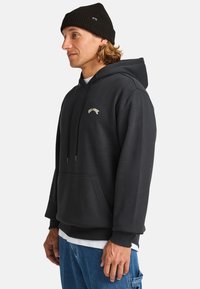 Felpa nera con cappuccio realizzata in morbido tessuto, dotata di una tasca frontale e di un logo ricamato in oro. Indossata con jeans blu e un berretto nero.