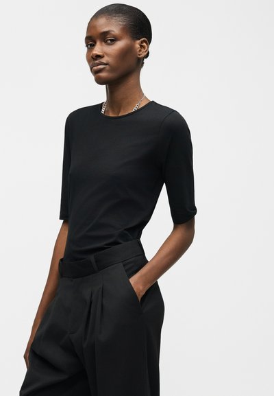 Γυναίκα που φοράει στενή μαύρη κοντομάνικη μπλούζα και μαύρο πλισέ παντελόνι ψηλόμεσο, με το ένα χέρι στην τσέπη, πάνω σε απλό φωτεινό φόντο.