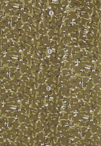 Gros plan d'une chemise à motif camouflage vert, marron et blanc, avec une poche avant et quatre boutons visibles.