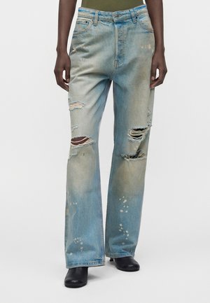 Person iført lyseblå slidte vidde jeans med flere rifter og malingsstænk, kombineret med sorte sko.