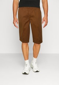 Hnědé kraťasy ke kolenům s plochým předním dílem, zapínáním na knoflík a poutky na opasek, v kombinaci s bílými ponožkami a teniskami. Hladká textura.