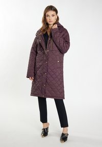 Palton lung cu captuseala de culoare burgundă, cu glug�ă, fermoar și nasturi cu capse, și buzunare laterale; asortat cu pantaloni negri și pantofi cu toc.