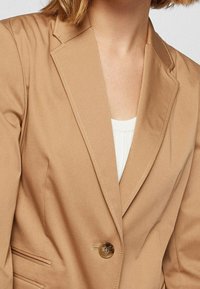 Kobieta w beżowej marynarce z klapami i jednym brązowym guzikiem, nosząca białą bluzkę, skróconą na szyi i w połowie torsu.