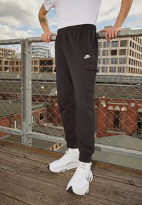 Svarta träningsoveraller med sidoficka och elastisk midja, ihop med vita sportskor. Ytan är slät, tyget ser mjukt ut.