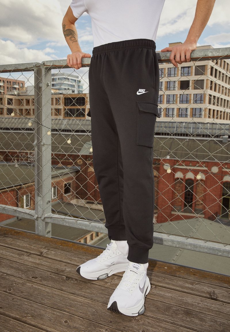 Svarta träningsoveraller med sidoficka och elastisk midja, ihop med vita sportskor. Ytan är slät, tyget ser mjukt ut.