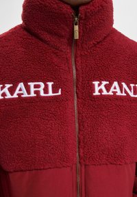 Sarkanais jaku ar rāvējslēdzēju, kuram ir pūkaina, fakturēta augšdaļa ar baltu izšuvumu "KARL"; krāsa pāriet uz gludāku apakšdaļu.