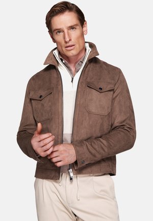 Man wearing a brown suede jacket over a white half-zip sweater and beige pants, adjusting his jacket sleeves against a plain background.