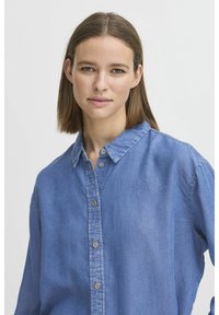 Camicia di denim azzurro chiaro con bottone davanti, colletto e maniche lunghe. Il tessuto ha una texture liscia e una leggera lucentezza.
