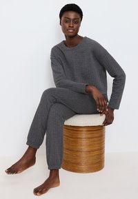 Graues gesteppte Lounge-Outfit mit langen Ärmeln und Bündchen. Modell sitzt auf einem runden Holzschemel mit einem strukturierten Stoffbezug.